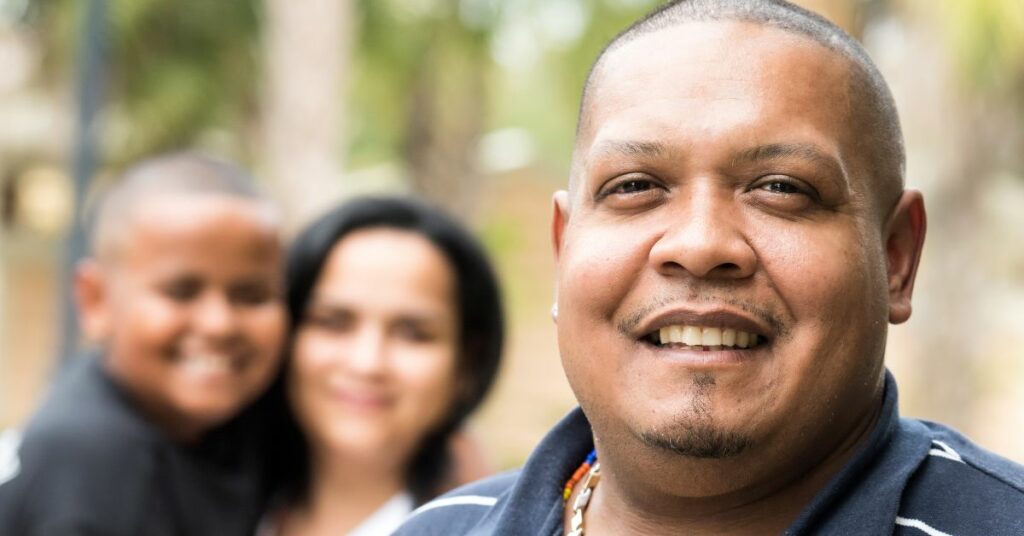 Close-up portrait of a man smiling in the foreground with a blurred background featuring a woman and child, conveying a sense of family and connection