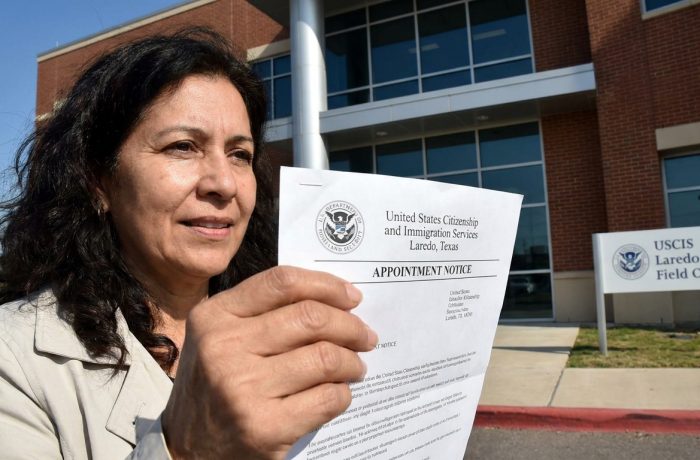 Exterior de la oficina de USCIS Laredo TX en Maher Avenue, con una persona esperando afuera sosteniendo una notificacion de cita.
